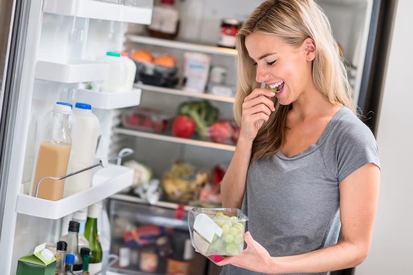 woman standing in front of an open refrigerator eating grapes