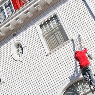 person climbing a ladder against a home exterior with paint can